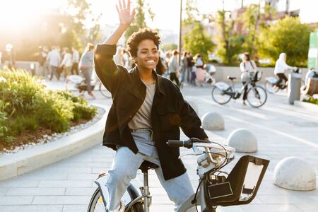 Photo of cute african american woman wearing casual clothes smiling and waving hand while sitting on bicycle in city parkの写真素材