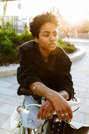 Photo of brooding african american woman wearing casual clothes looking aside while sitting on bicycle in city parkの写真素材