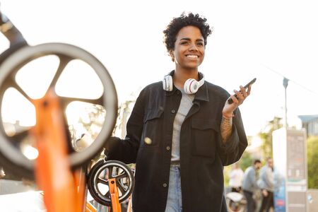 Photo of beautiful african american woman standing with headphones and cellphone near parking for...