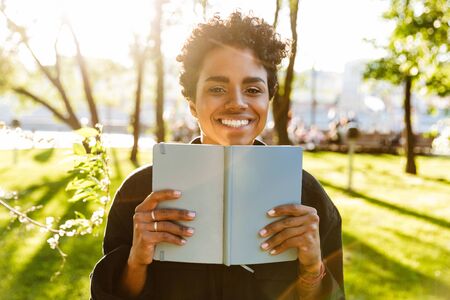 Photo of adorable african american woman with curly hair smiling and holding day planner while walking in city parkの写真素材