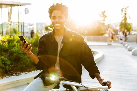 Photo of pleased african american woman wearing casual clothes smiling and holding cellphone while sitting on bicycle in city parkの写真素材