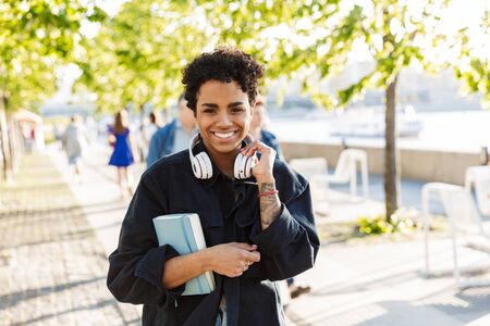 Portrait closeup of pleased african american woman with curly afro hairstyle holding diary and headphones while walking along riverfrontの写真素材