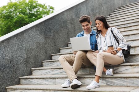 Image of optimistic couple man and woman with paper cup smiling and looking at laptop while sitting on stairs outdoorsの写真素材