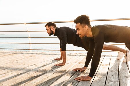 Two attractive young healthy sportsmen outdoors at the beach, workout together, plank exerciseの写真素材