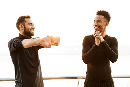 Two attractive smiling young healthy sportsmen outdoors at the beach, workout together, doing stretching exercisesの写真素材