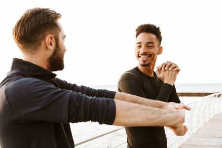 Two attractive smiling young healthy sportsmen outdoors at the beach, workout together, doing stretching exercisesの写真素材