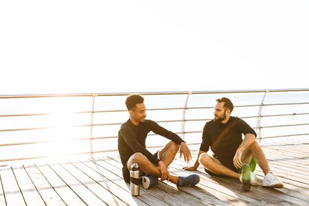 Two attractive smiling young healthy sportsmen sitting outdoors at the beach, resting after jogの写真素材