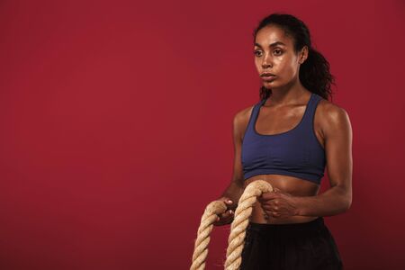 Photo of a concentrated strong young african fitness sports woman posing isolated over red wall background posing with ropes.の写真素材