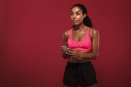 Photo of a pleased smiling positive strong young african fitness sports woman posing isolated over red wall background using mobile phone chatting.の写真素材