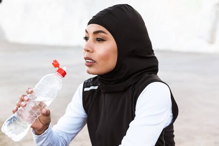 Attractive muslim sportswoman wearing hijab outdoors, sitting, drinking water