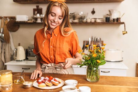 Picture of a pleased smiling cheery young blonde girl chef cooking at the kitchen strawberry sweet pastry.の写真素材
