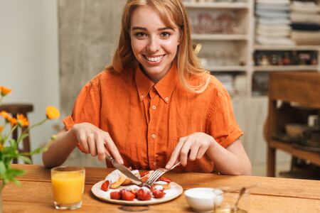 Image of a happy young blonde girl at the kitchen eat sweet pastry have a breakfast drinking juice.の写真素材