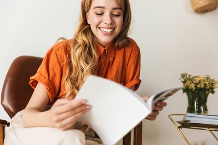 Image of a pleased smiling young blonde girl at home indoors reading magazine sitting on chair.の写真素材
