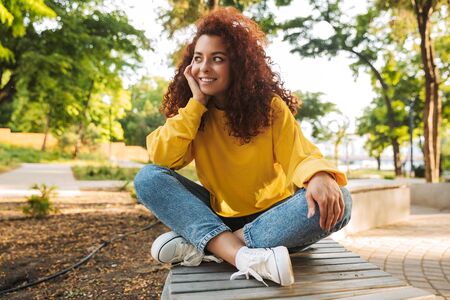 Photo of a smiling cheerful happy young beautiful curly student girl outdoors in nature park sitting on a bench.の写真素材