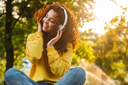 Beautiful young cheerful girl spending good time at the park, sitting on a bench, listening to music with headphonesの写真素材