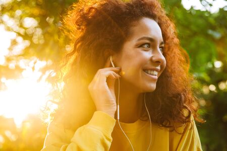 Image of a happy optimistic cute young student curly girl sitting on bench outdoors in nature park with beautiful sunlight listening music with earphones.の写真素材