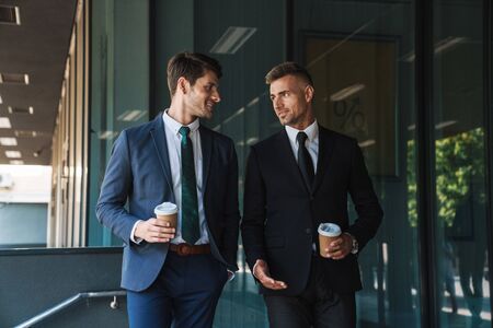 Image of caucasian businessmen partners dressed in formal suit talking and drinking takeaway coffee while standing outside office centerの写真素材