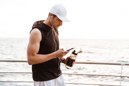 Photo closeup of handsome man in white cap using smartphone and drinking water while walking on pier at seaside in morningの写真素材
