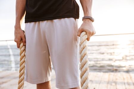 Photo cropped of caucasian man in tracksuit holding fitness rope on wooden pier at seaside in morningの写真素材