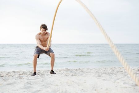 Photo of brutal half-naked man working out with fitness rope on beach at seaside in morningの写真素材