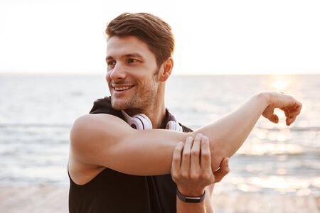 Photo smiling man in tracksuit with headphones doing exercise while working out at seaside in morningの写真素材