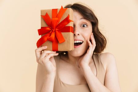 Portrait of optimistic caucasian woman 20s dressed in casual clothes smiling at camera while holding present box isolated over beige background in studioの写真素材