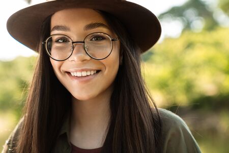 Photo of joyful young woman with long dark hair wearing stylish hat and eyeglasses walking in green park on sunny dayの写真素材