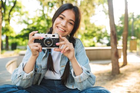Photo of a cheerful optimistic cute young student girl wearing eyeglasses sitting on bench outdoors in nature park holding camera photographing.の写真素材