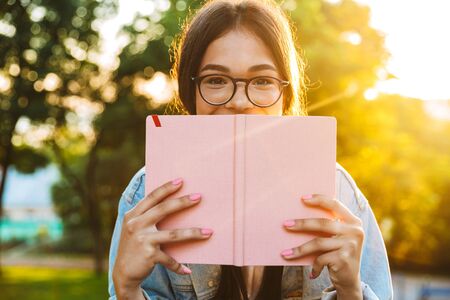 Image of a pretty cute young student girl wearing eyeglasses sitting outdoors in nature park covering face with book.の写真素材