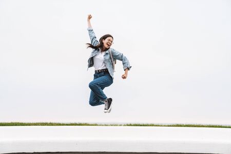 Image of cheerful teenage girl dressed in casual wear having fun and rejoicing while walking along white wall outdoorsの写真素材