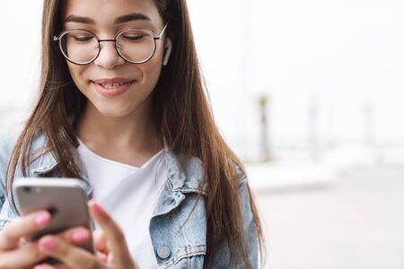 Image of adorable smiling teenage girl wearing earpods and eyeglasses using smartphone while walking outdoorsの写真素材