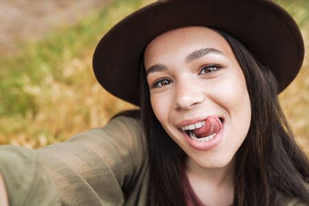 Photo closeup of cheerful woman with long dark hair wearing hat showing her tongue at camera while taking selfie photo outdoorsの写真素材