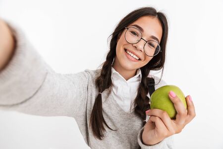Image of happy young school girl isolated over white wall background posing take a selfie by camera eat apple.の写真素材