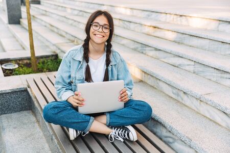 Photo of a smiling pleased cute young student girl wearing eyeglasses sitting on bench outdoors in nature park using laptop computer.の写真素材