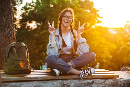 Photo of a cheerful cute young student girl wearing eyeglasses sitting on bench outdoors in nature park with beautiful sunlight showing peace gesture.の写真素材
