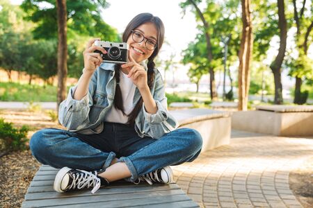 Photo of optimistic cheerful happy cute young student girl wearing eyeglasses sitting on bench outdoors in nature park holding camera photographing.の写真素材