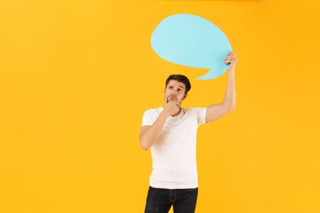 Photo of serious man in basic t-shirt thinking and holding blank thought bubble isolated over yellow...