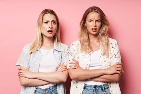 Two shocked attractive blonde girls wearing summer outfit standing isolated over pink background