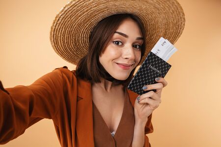 Beautiful young woman wearing straw hat and summer outfit standing isolated over beige background, taking a selfie while holding passport with flight ticketsの写真素材