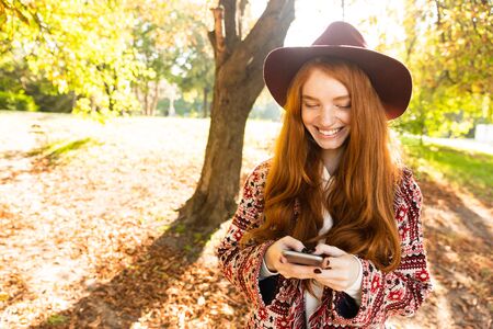 Image of a smiling cutie young student redhead girl in autumn park using mobile phone.の写真素材