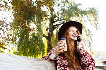 Image of a positive pleased happy cheery young student redhead girl in autumn park talking by mobile phone drinking coffee.の写真素材