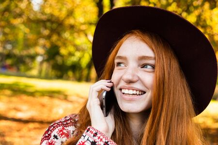 Picture of a smiling pleased cutie young student redhead girl in autumn park talking by mobile phone.の写真素材