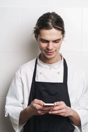 Image of a handsome young cook chef at the kitchen using mobile phone chatting.の写真素材