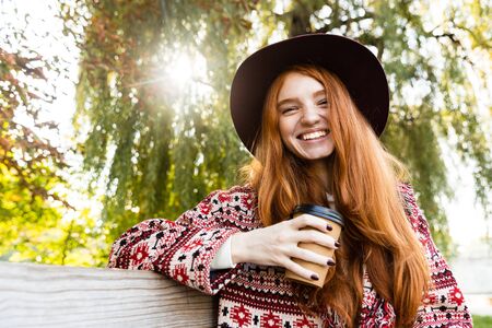 Image of a positive pleased young student redhead girl in autumn park drinking coffee.の写真素材