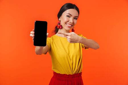 Image of a beautiful excited happy young woman posing isolated over orange wall background showing display of mobile phone.の写真素材