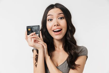 Image of cheerful brunette woman 20s dressed in basic clothes smiling and holding credit card...