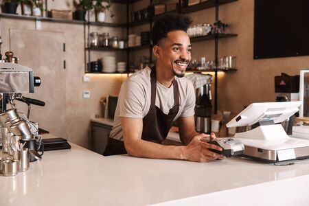 Smiling attractive african man barista standing behind the counter at the coffee shopの写真素材