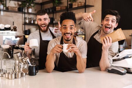 Group of cheerful men baristas wearing aprons working at the counter in cafe indoorsの写真素材