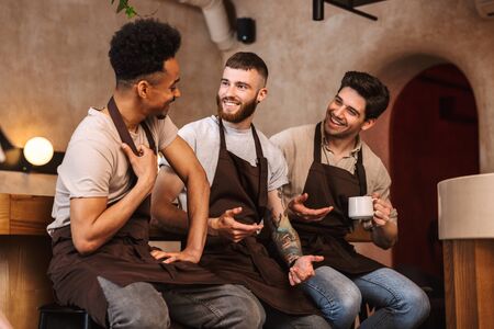Three cheerful male baristas standing at the coffee shop counter indoorsの写真素材