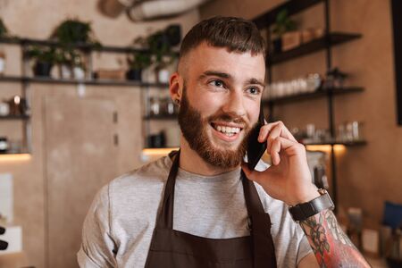 Happy young man barista standing at the coffee shop, talking on mobile phoneの写真素材